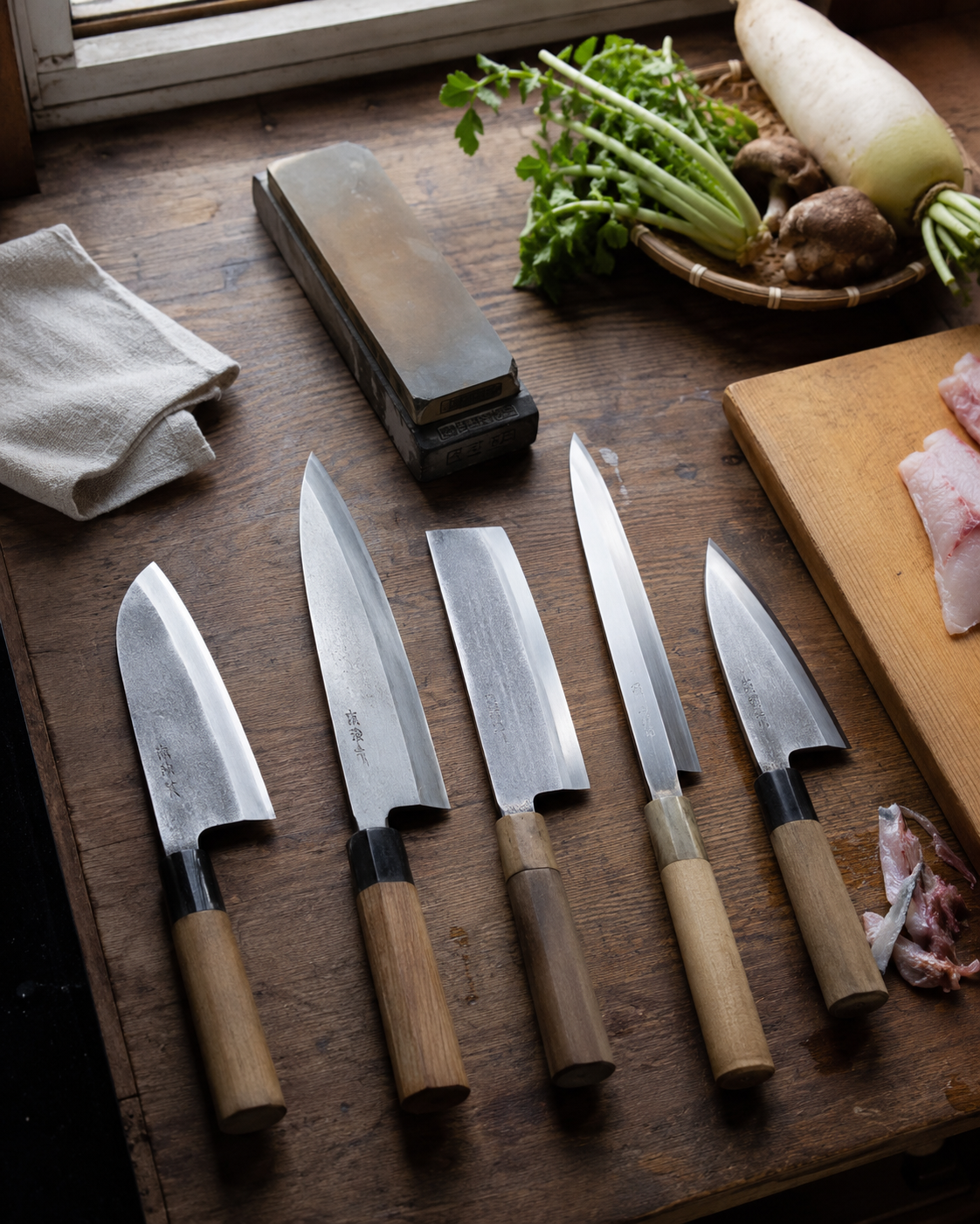 Professional Japanese chef knives and whetstone on a rustic wooden kitchen counter with fresh vegetables.